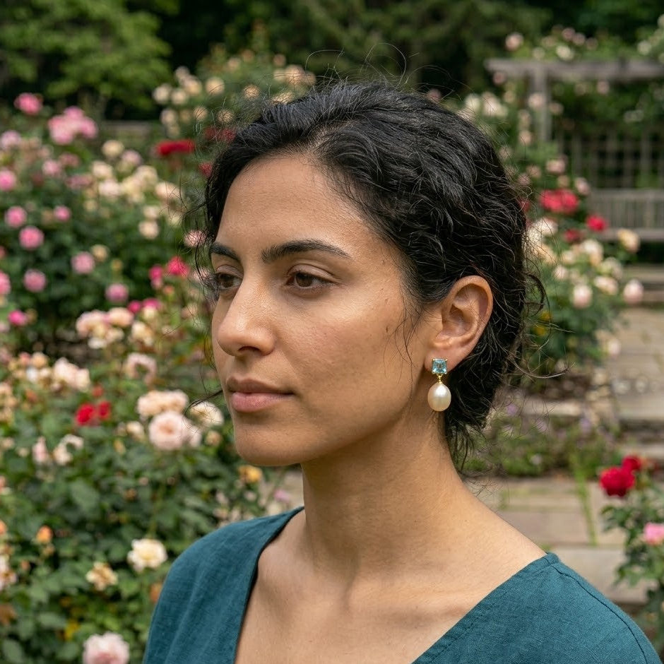 Woman with dark hair and earrings standing in a garden with flowers and steps in the background