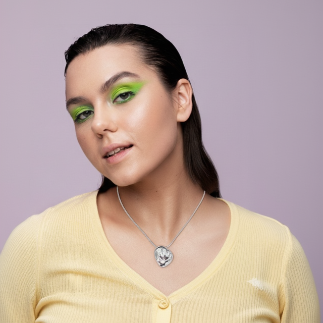 Woman wearing a yellow top and a necklace with a heart pendant against a light purple background