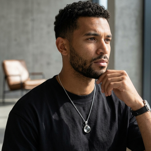 Man wearing a black shirt and necklace, sitting indoors with a blurred background