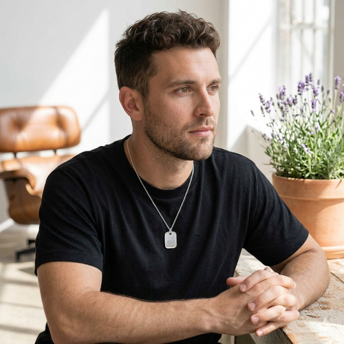 Man wearing a black t-shirt with a necklace, sitting indoors with a plant in the background
