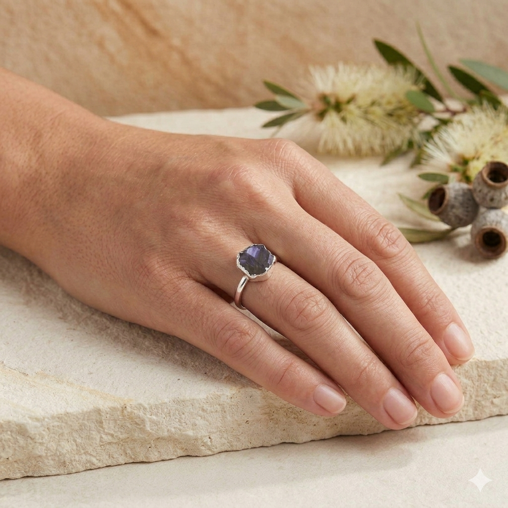 Hand wearing a ring with a blue gemstone on a natural stone surface with flowers in the background