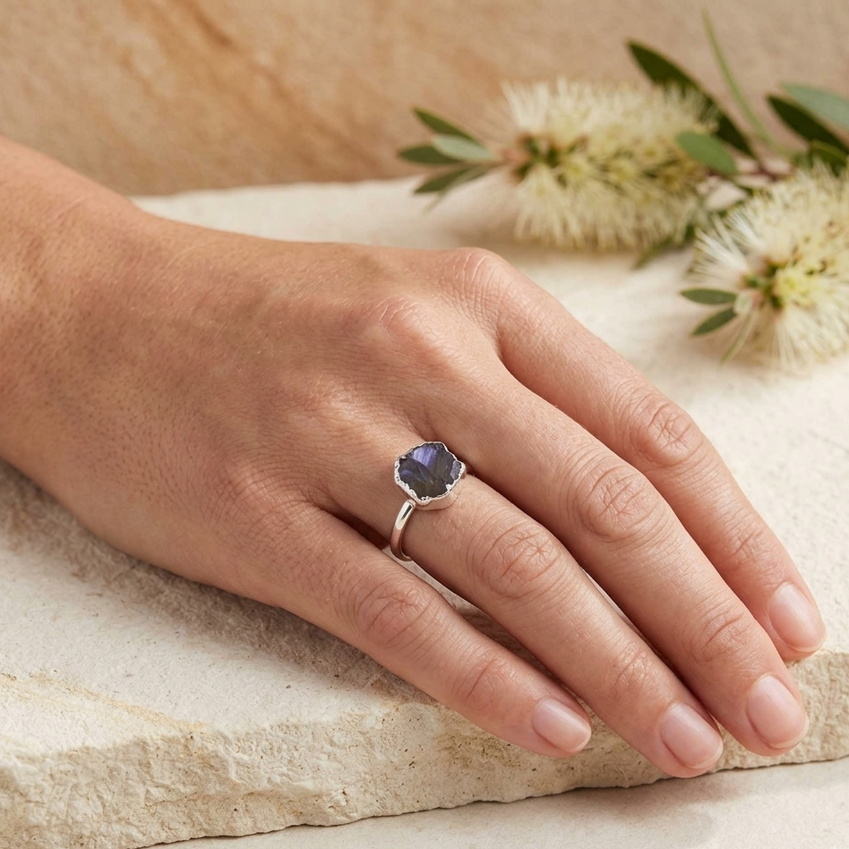 Hand wearing a ring with a blue gemstone on a stone surface with flowers in the background
