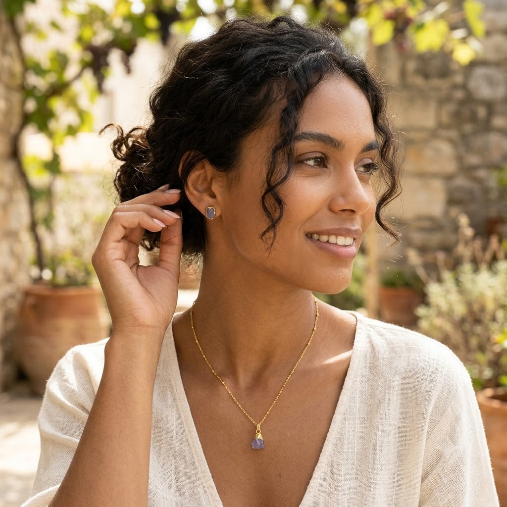 Woman wearing a gold necklace and earrings outdoors with plants and stone wall in the background